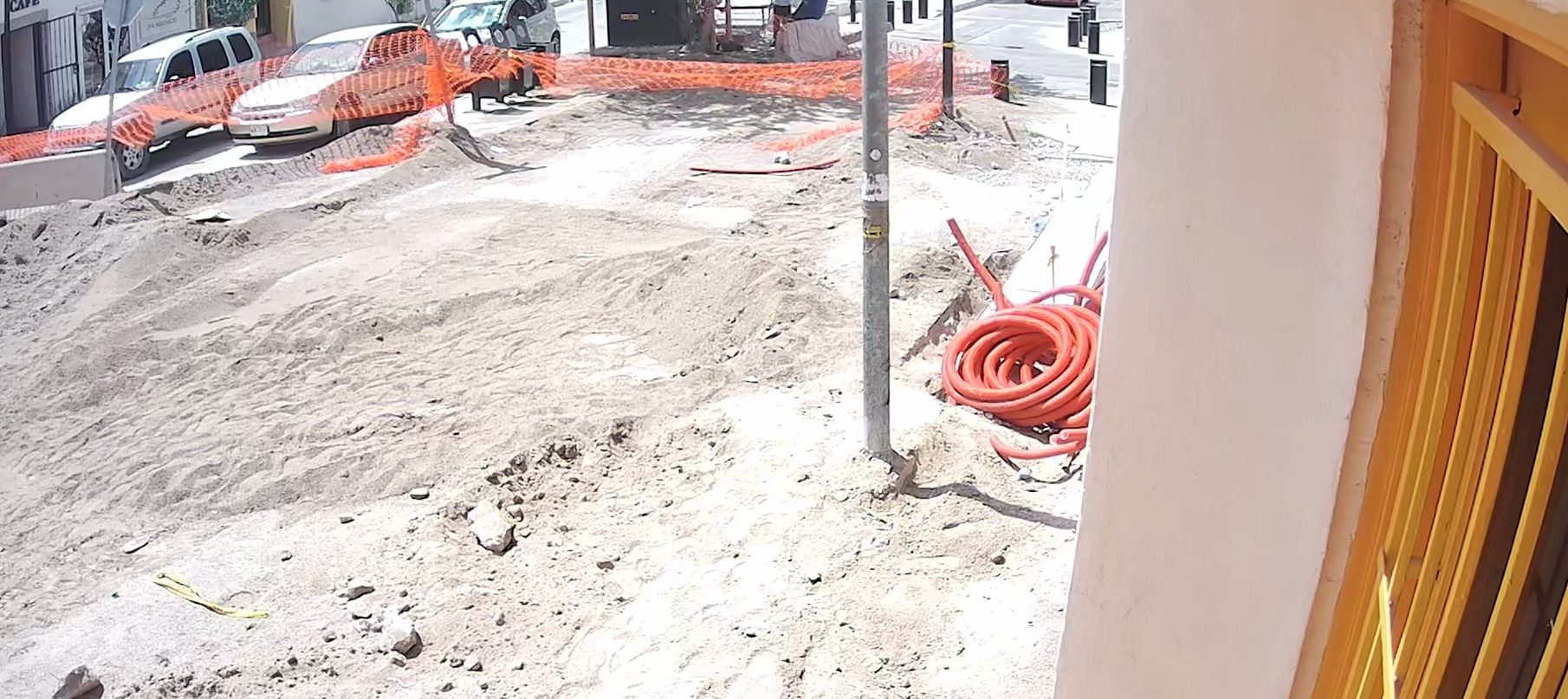 La calle frente al Tiny Room en obra: terracería, malla naranja y rollos de tubo; a la derecha, la puerta amarilla